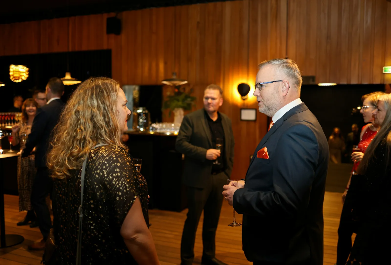 Senior professionals in conversation at an evening business event, drinks in hand, in a warmly lit wood-paneled room